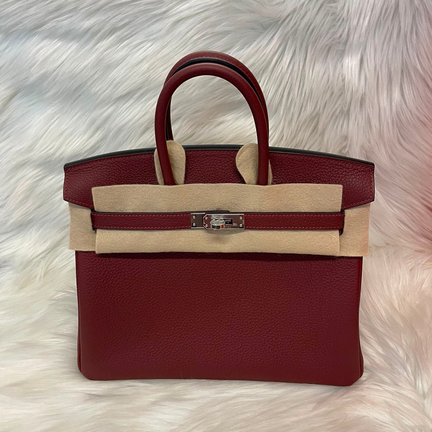 A structured leather handbag in deep burgundy with beige canvas trim and a silver-toned front clasp, displayed on a white faux fur surface.