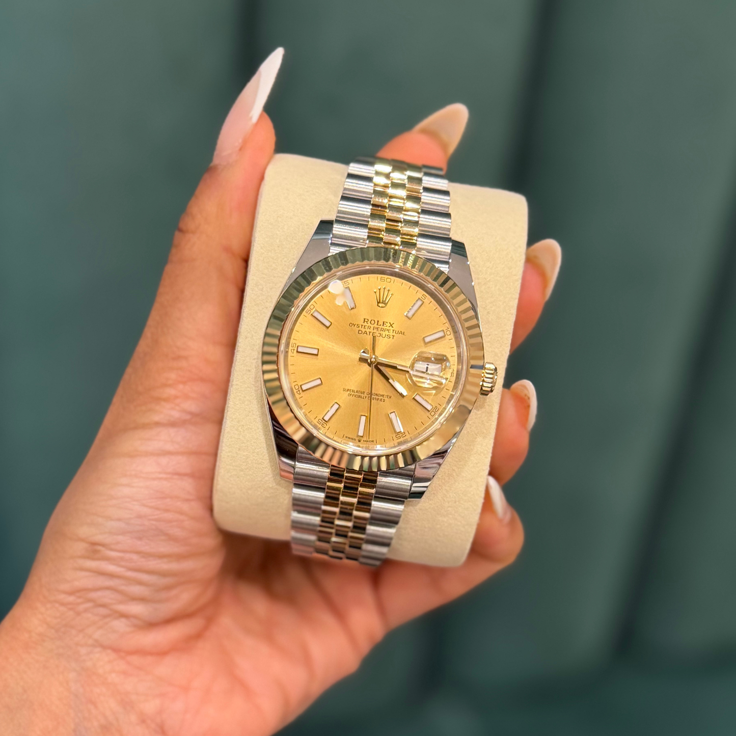 A Rolex Datejust watch with a gold dial and two-tone gold and silver bracelet, held in a hand against a dark green background.