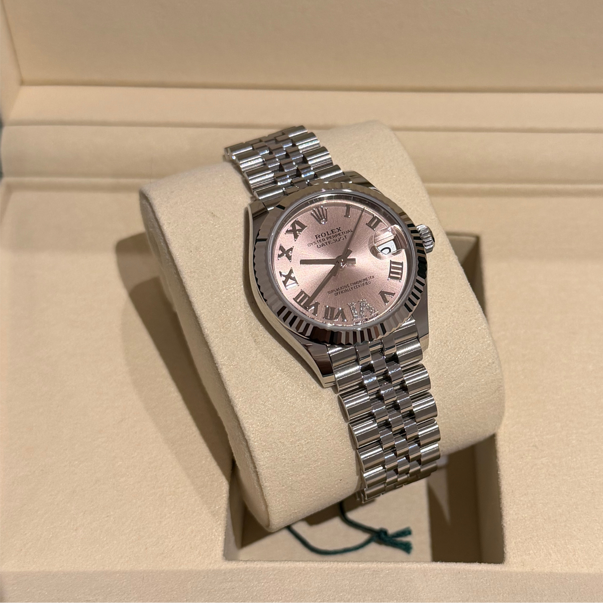 A Rolex Datejust watch with a silver-tone metal bracelet and a pink dial, displayed on a beige watch cushion inside its presentation box.