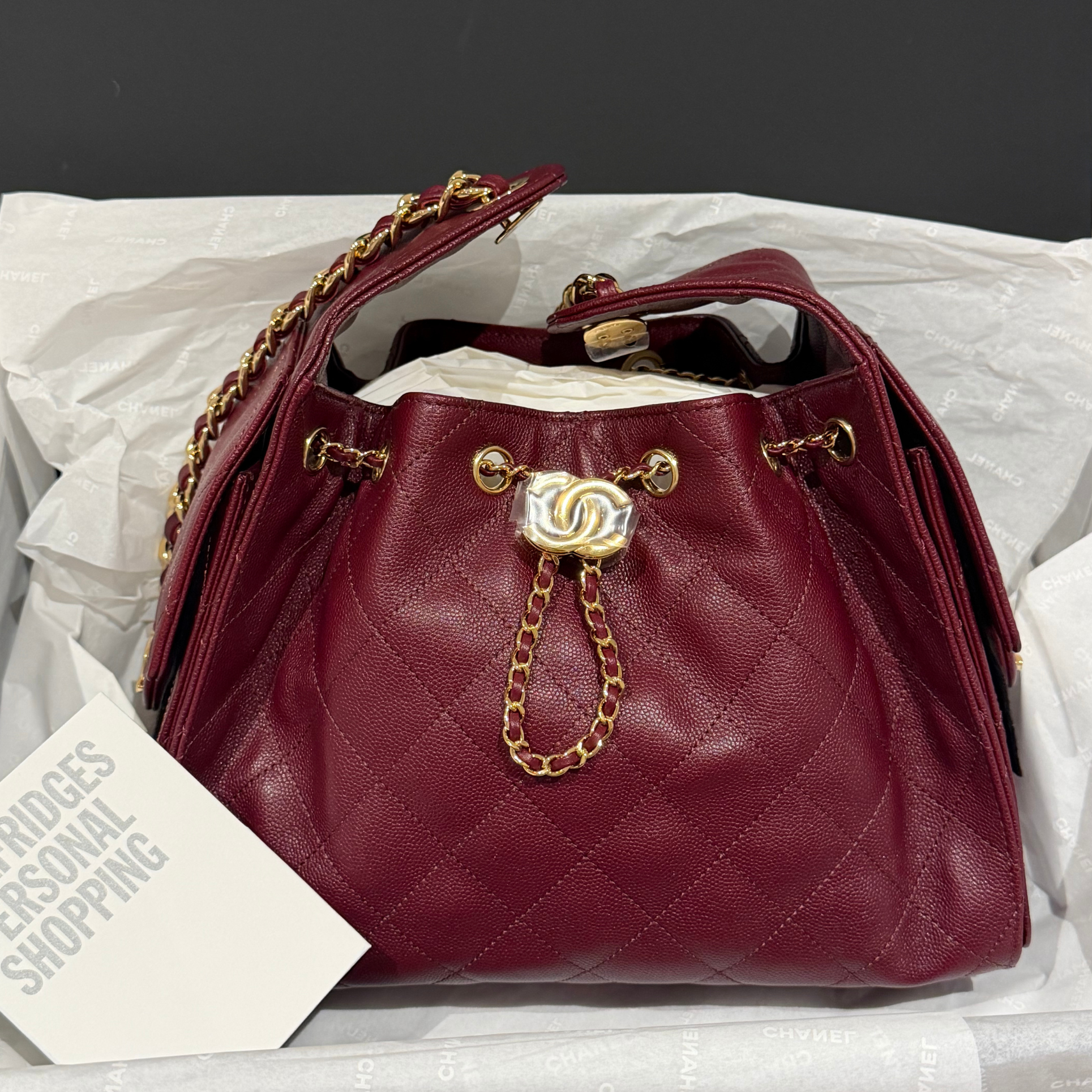 A burgundy quilted leather handbag with gold-tone chain strap and hardware, displayed in its white branded packaging with a card reading CHANEL'S PERSONAL SHOPPING.
