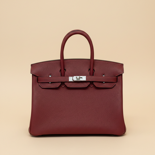 A structured leather handbag in deep burgundy with a wide beige fabric bow detail across the front and a silver-toned clasp, photographed against a white faux-fur surface.
