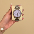 A silver-tone stainless steel wristwatch with a pale pink dial and matching pink hour markers, held in a hand on a cream-colored display block. The watch is shown in a lifestyle setting with a neutral gray background.