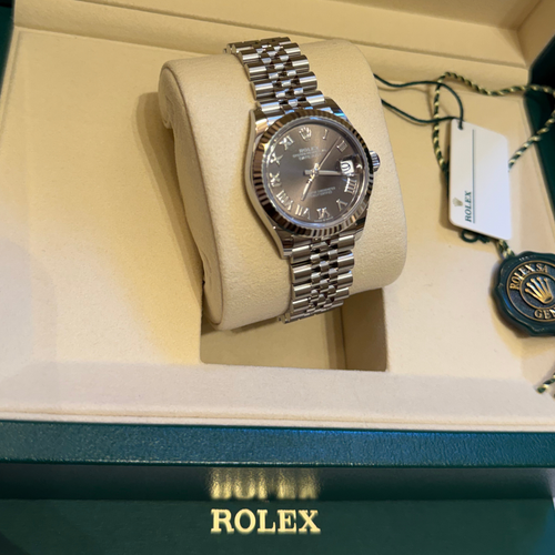 A Rolex Datejust watch with a stainless steel bracelet and dark gray dial, displayed inside a cream-colored Rolex gift box with its original packaging tags visible.