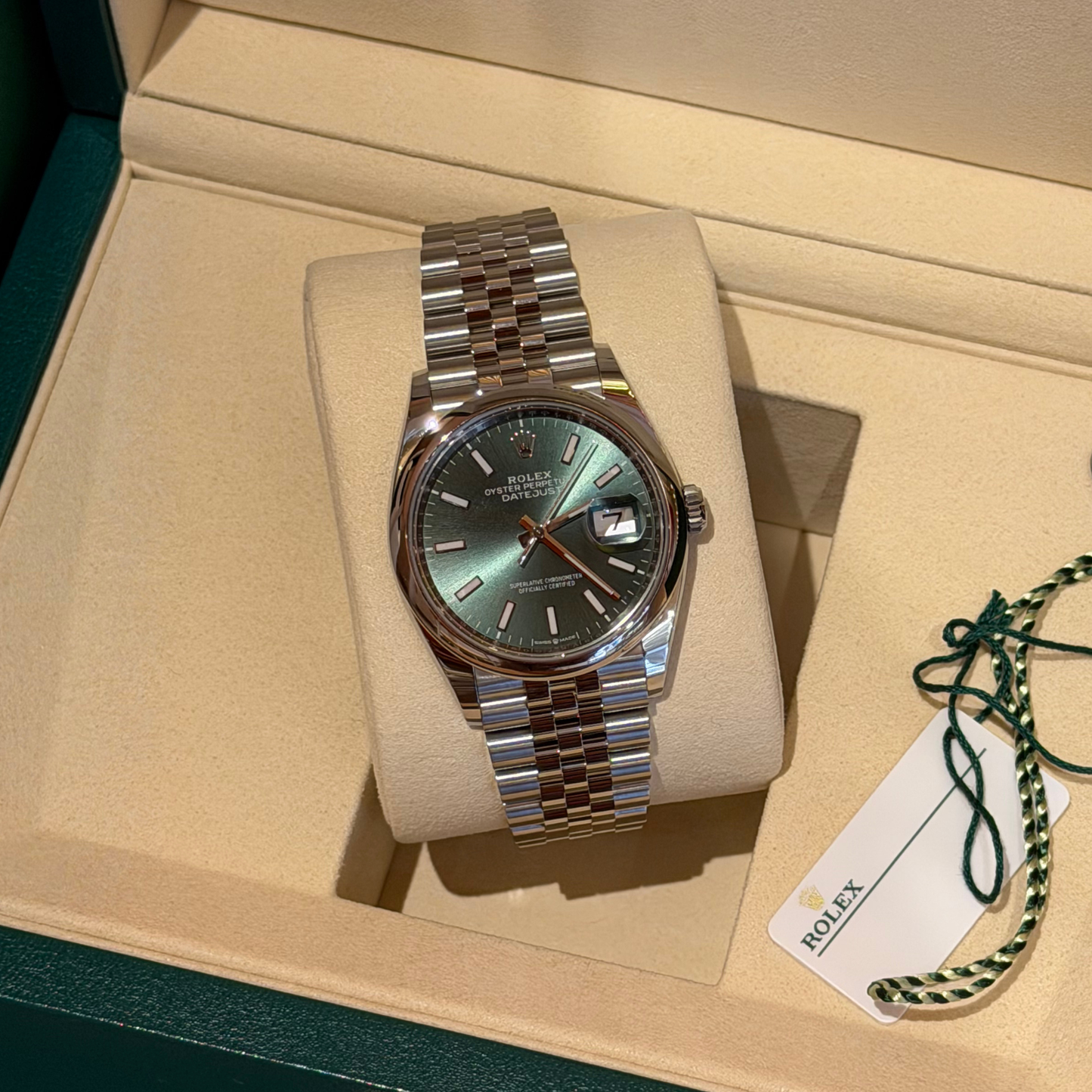 Rolex Datejust watch with a green dial and two-tone stainless steel Oyster bracelet, displayed on a cream cushion inside its original box with a green string and paper tag.