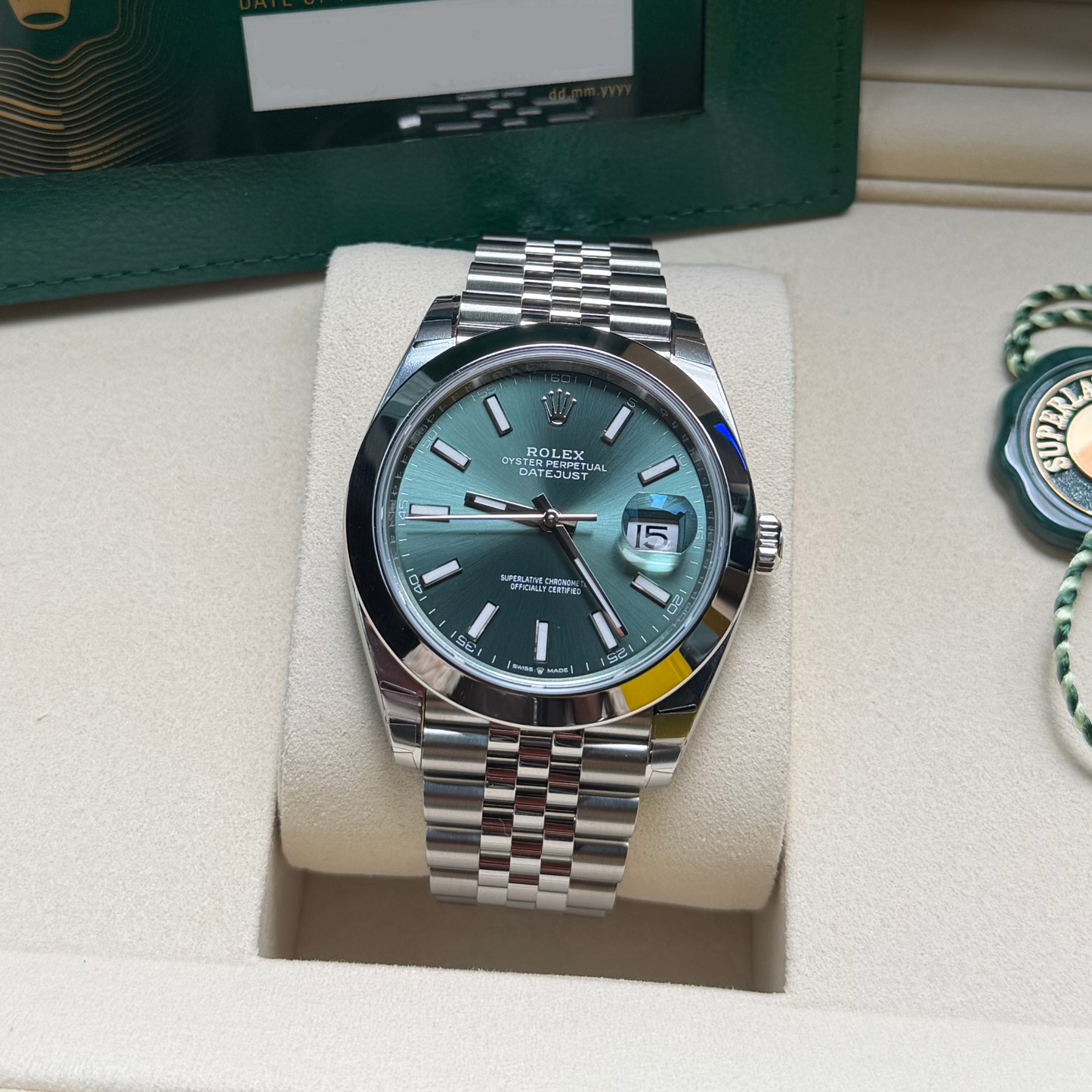 Rolex Datejust wristwatch with a green dial and stainless steel Oyster bracelet, displayed on a cream cushion inside a watch box.