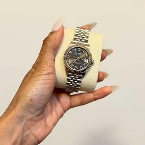 A silver stainless steel wristwatch with a dark gray dial and matching steel Oyster bracelet, held on a cream-colored display cushion by a hand with long nude manicured nails.