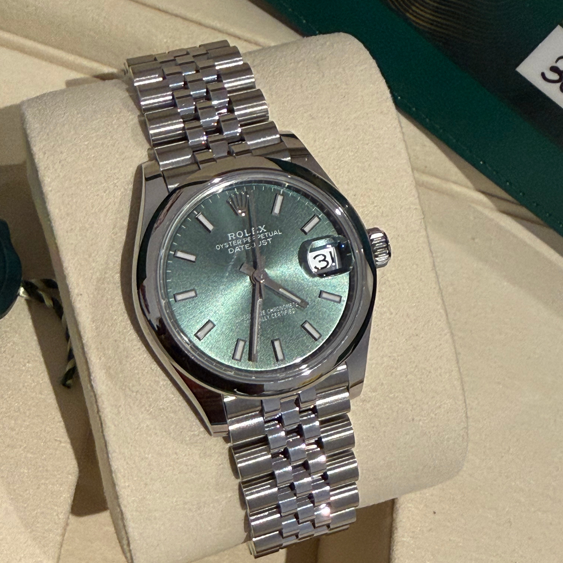 A Rolex Datejust watch with a green dial and a silver Oyster bracelet, displayed on a beige cushion inside its box.