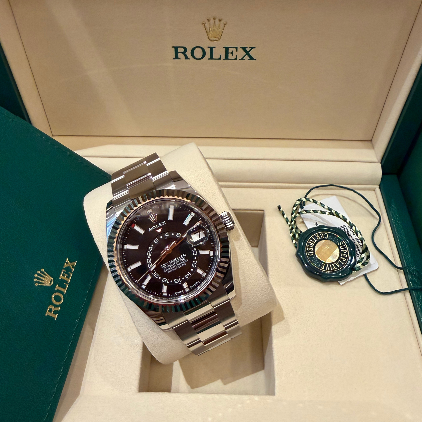 A Rolex wristwatch displayed inside an open Rolex presentation box, with the watch on a cream cushion and a small branded tag beside it. The watch has a dark brown dial, stainless steel bracelet, and fluted bezel.