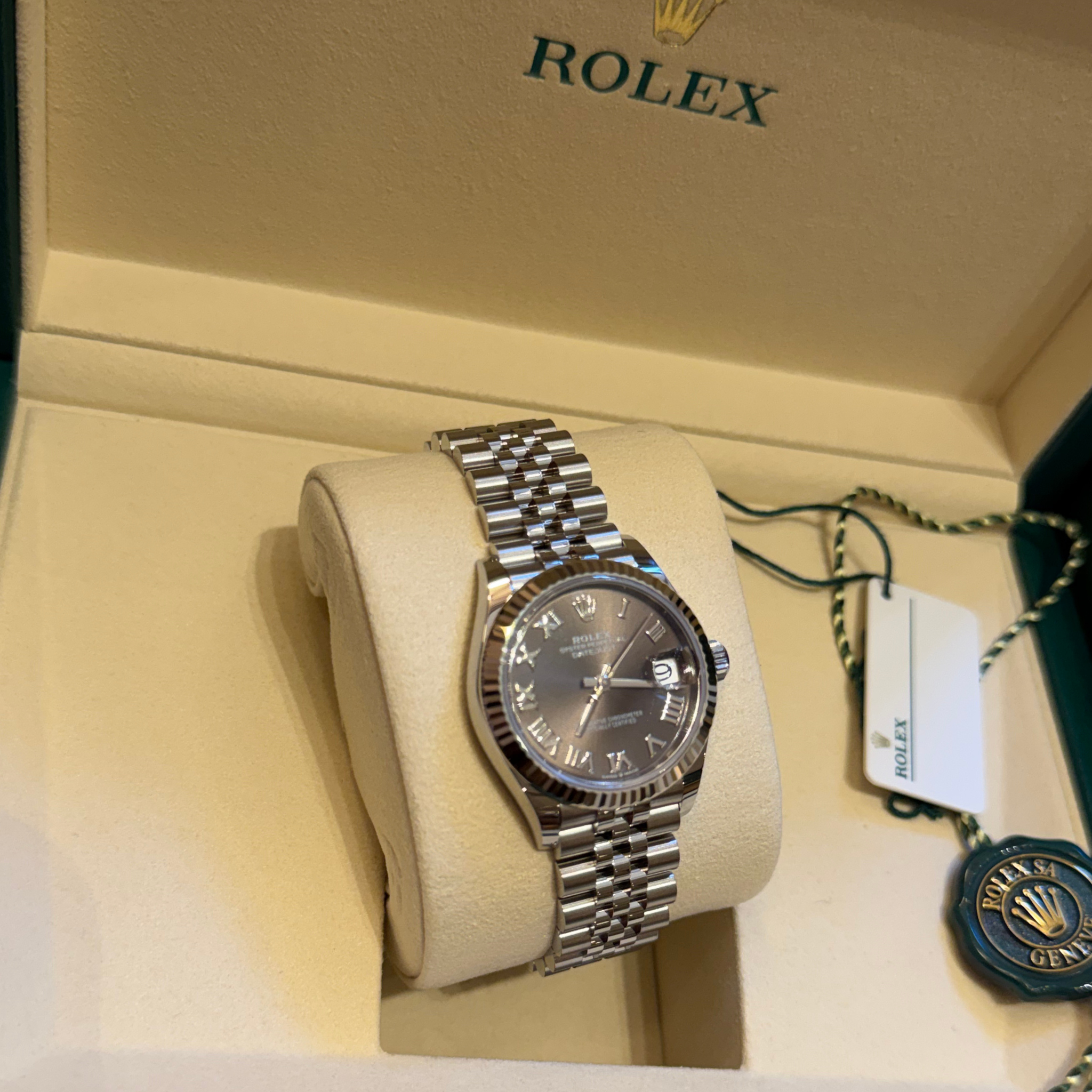 Rolex wristwatch displayed on a cream-colored Rolex presentation cushion inside its original box, with the watch featuring a silver-tone stainless steel Oyster bracelet and a dark gray dial.