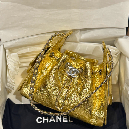 A metallic gold Chanel bucket bag with a silver-tone interlocking CC logo and chain strap, resting on a black Chanel dust bag inside its white packaging box.