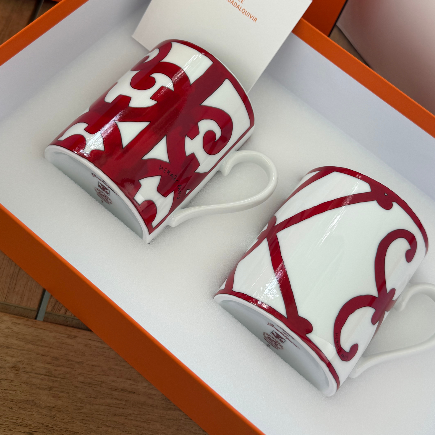 Two white ceramic mugs with red ornamental scrollwork patterns, presented inside an orange Hermes box with a white insert card.
